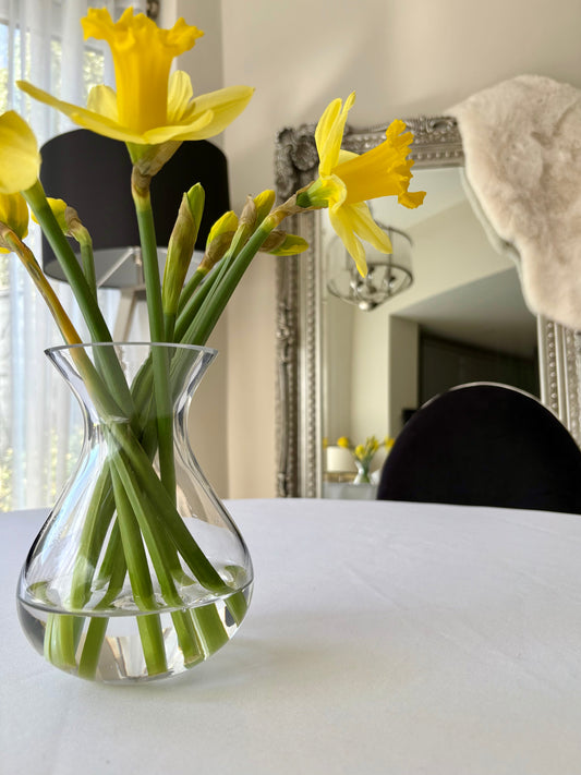 White tablecloth on a dining table with a small clear vase of bright yellow daffodils, creating a simple and elegant spring tablescape.